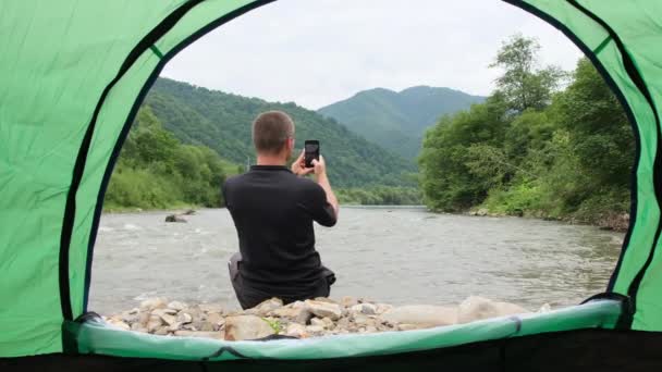 荒れ狂う川のほとりにあるテントからの美しい景色で 若い男が座ってスマートフォンで写真を撮っています 山々を眺めながら美しい写真を撮る — ストック動画