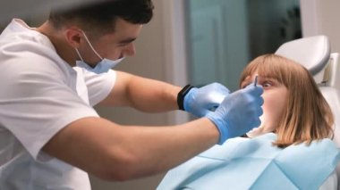 Korkmuş bir kız dişçi koltuğunda oturur. Korkmuş ve korkmuş görünüyor. Yetişkinlerin elinde lateks eldivenler dişçi aletleri var..