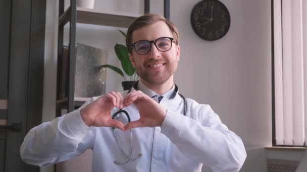 의사는 심장의 몸짓을 주면서 카메라를 보면서 웃는다 사람들을 한다는 — 비디오