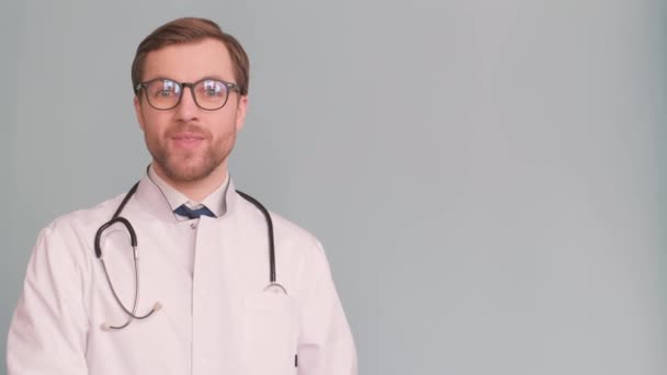 灰色の背景に隔離された彼の手で何かを示し 提示する若い男性医師の笑顔 高品質4Kビデオ — ストック動画