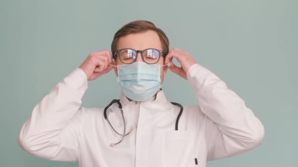医者の肖像画は 灰色の背景に隔離された保護マスクを脱いで 彼はカメラを見ている 4Kビデオ — ストック動画