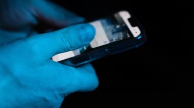 Bir adamın eli parmaklarını hızlı bir şekilde akıllı telefonun ekranında hareket ettirir ve internette bir şeyler arar. Mavi ışıkta akıllı telefonu olan bir erkek.
