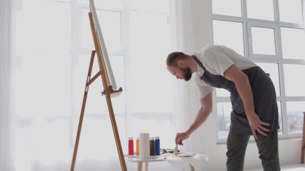 筆を使って才能ある男性アーティストが 白いキャンバスに油彩画の現代的な傑作を作成します キャンバスは大きなスタジオでイーゼルの上にあります — ストック動画