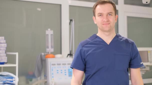 診療所の事務所に腕を組んで立っている青いコートのハンサムな医者の肖像画 自信に満ちた医者がカメラを見て微笑んだ 4Kビデオ — ストック動画