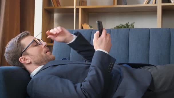 オフィスのラウンジエリアに横たわっている間 若いビジネスマンがスマートフォンで話しています 仕事の疲れ 業務の呼び出しの概念 — ストック動画