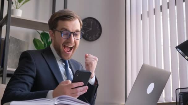 オフィスで手にスマートフォンを持つ面白いビジネスマンは幸せとエネルギーを感じている 幸福感の状態でクレイジーオフィスの労働者 — ストック動画