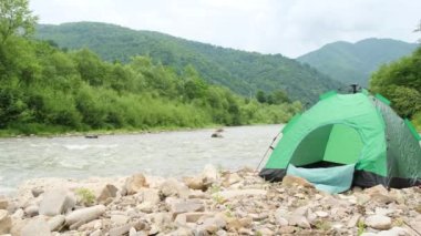 Bulutlu havada dağlık arazinin güzel yaz manzarası. Dağ nehrinin yanında yeşil bir çadır. Ekolojik turizm kavramı