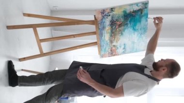 Yetenekli bir sanatçının beyaz bir tuvalde modern bir sanat eseri yaratmak için fırça kullanmasının dikey videosu. Tuval atölyede bir sehpanın üzerinde..