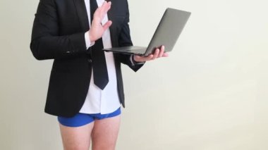 Pantolonu olmayan iş ceketli bir adam dizüstü bilgisayar tutuyor ve internette biriyle konuşuyor. Beyaz bir duvarın karşısında iç çamaşırlı bir iş adamı..