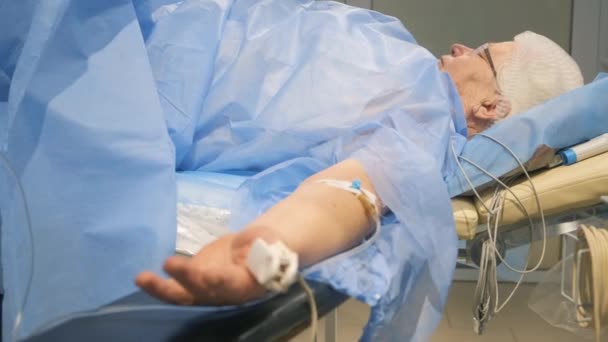 古い白髪の祖父が手術中に手術台の上に横たわっています チューブで接続された注射で患者の腕 — ストック動画