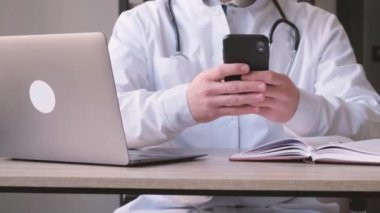 Erkek bir doktor elinde bir akıllı telefon tutuyor ve klinikteki bir çalışma masasında oturuyor. Profesyonel bir doktorun internet danışmanlığı. Tıp kavramı