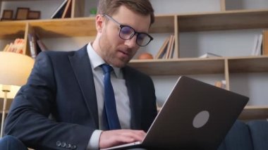 Genç başarılı iş adamı resmi iş süitinde sakallı, koltukta, lüks ofis içinde dizüstü bilgisayarla oturuyor..