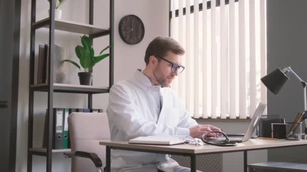 オフィスのテーブルに座り白い医療用の制服を着たセラピストとして働いています — ストック動画