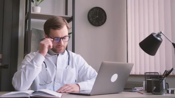 家族の医者はノートパソコンの診療所に座って眼鏡を外してカメラを見て笑う — ストック動画