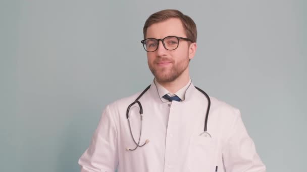 一个戴着灰色背景眼镜的年轻医生的画像 他看着镜头 挥挥手 治疗人的概念 — 图库视频影像