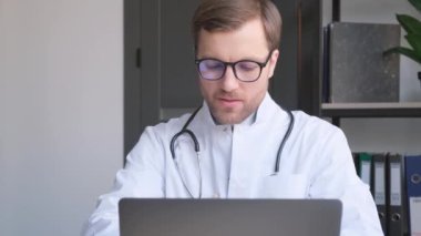 Genç yakışıklı bir doktor ofisinde bir dizüstü bilgisayar üzerinde çalışıyor. Avrupa 'da modern bir sağlık kliniği.