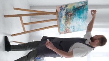 Yetenekli bir sanatçının beyaz bir tuvalde modern bir sanat eseri yaratmak için fırça kullanmasının dikey videosu. Tuval atölyede bir sehpanın üzerinde..