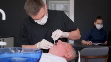 Erkek bir dişçi, dişçilik konsepti olan klinikte dişlerini inceleyen yaşlı bir adamla çalışıyor. Diş implantlarının yaşlılıkta yerleştirilmesi. Yüksek kalite 4k video