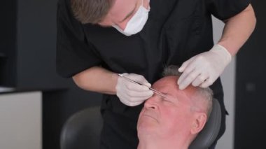 Cerrah, estetik ameliyattan önce yaşlı bir adamın yüzünü inceleyerek göz bölgesini değiştirir. Doktor estetik ameliyata hazırlanıyor..