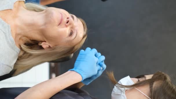 美容クリニックで注射を行う美容師の垂直ビデオ 医者は老女の額に抗老化薬を注射する — ストック動画