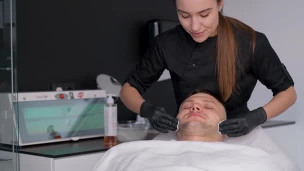 顔の治療を受けている温泉の若い男 人の顔に粘土マスク 美容師が顔につけられた保湿マスクで顔のマッサージを行います — ストック動画