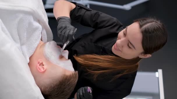 女性美容師による洗顔の最中のハンサムな男 粘土マスクを若返らせる 垂直ビデオ — ストック動画