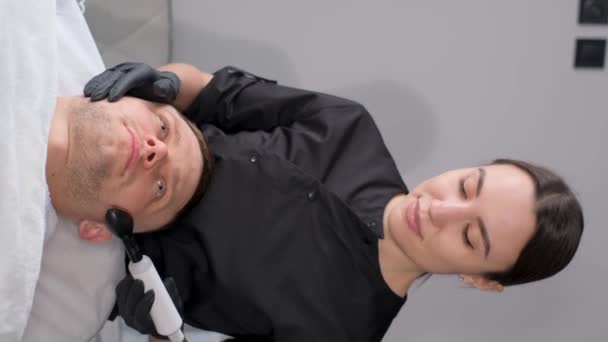 一位美容师在一位年轻英俊男性客户的脸上使用射频升降装置拍摄的垂直视频 温泉美容院 — 图库视频影像