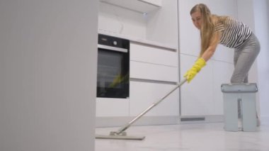 Vücudunun tüm görüntüsü, güzel, neşeli, çalışkan bir kız. Hızlı ev işleri yapıyor, modern açık beyaz mutfağın kusursuz seramik zeminini temizliyor..