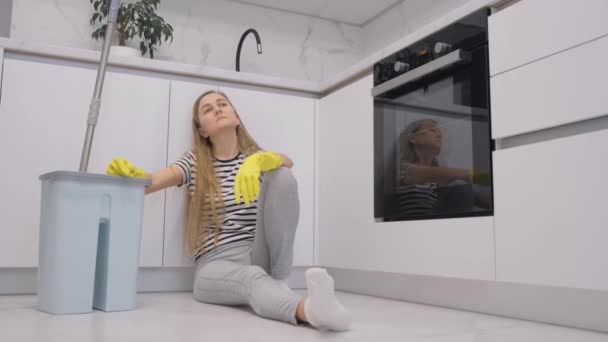 家を掃除した後 台所で休んでいる疲れた若い掃除の女性 バケツと掃除のためのモップで床に座って — ストック動画