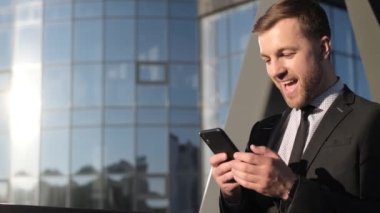İş merkezinin yanında elinde cep telefonu olan genç bir işadamı. Siyah takım elbiseli gülümseyen genç bir işadamı akıllı telefona bakıyor. Dünya iş konsepti