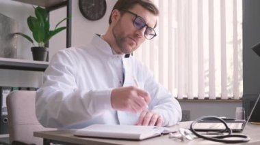 Gözlüklü ve üniformalı profesyonel genç doktor ofisinde evrak işleriyle uğraşıyor. Teşhis, tedavi planı yazıyorum. İş yerinde bir doktor..