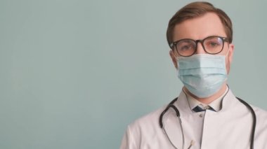 Tıbbi üniforma ve maskeyle mavi arka planda duran genç bir doktorun portresi. Ciddi odaklı doktor kameraya bakıyor.