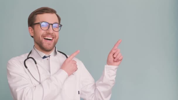 非常开心的笑着年轻的医生实习生摆出蓝色背景的相机 一个穿着白色医疗制服的有前途的年轻医生指着某人的广告镜头 — 图库视频影像