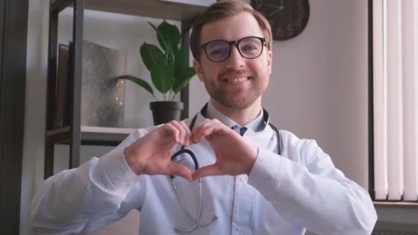在他的现代办公室里 一个年轻的欧洲型正正正医生站在镜头前微笑的画像 心脏病医生用自己的手做心脏动作 — 图库视频影像