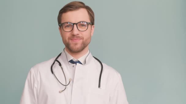 喜形于色 面带微笑的欧洲年轻医生 背景是蓝色的 一个有前途的年轻医生的广告拍摄 医生竖起大拇指 — 图库视频影像