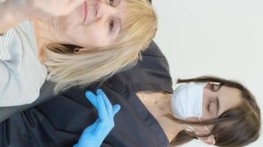 Tatmin olmuş yaşlı bir kadının trichologla olan randevusunun dikey videosu. Genç bir doktor hastaların kafa derilerini tarıyor.