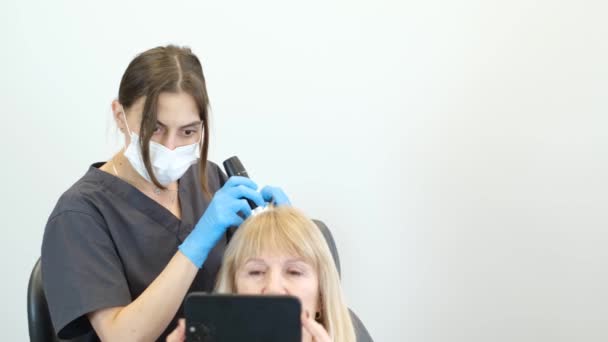 若いトリコロジストは高齢の患者の頭部を調べ 電話で放送する 頭皮のタイムリーな治療 — ストック動画