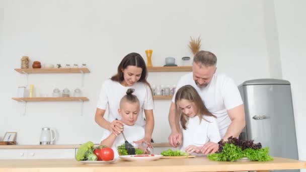 2人の小さな子供を持つ幸せな若い家族は 家庭のキッチンで一緒に料理を楽しみ 笑顔の親が小さな子供に健康的な食べ物 有機サラダ ディナーを調理する方法を教えています — ストック動画