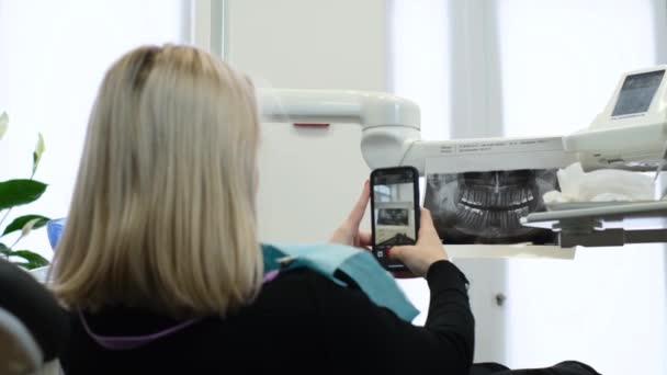 金髪の女性の患者は 歯の椅子に座っている間 スマートフォンで歯のX線を取ります 歯医者の若い患者 — ストック動画