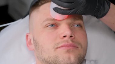 Elektronik masaj aleti kullanarak erkek yüz masajına yakın çekim. Bir kozmetik uzmanı genç sarışın bir adam için canlandırıcı yüz prosedürleri uyguluyor.