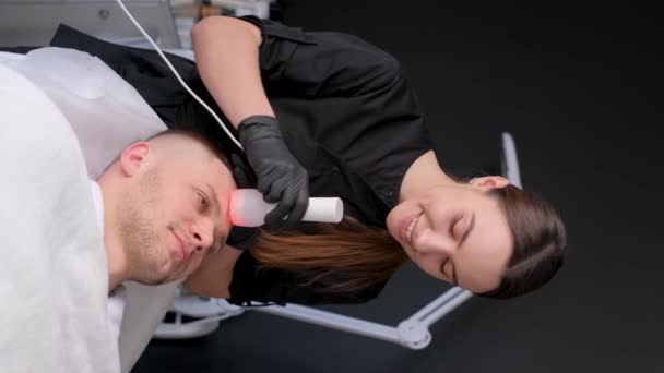 垂直录像 男性面部按摩使用电子多功能按摩器 一个美容师女孩为一个年轻貌美的男人做着年轻化的手术 — 图库视频影像
