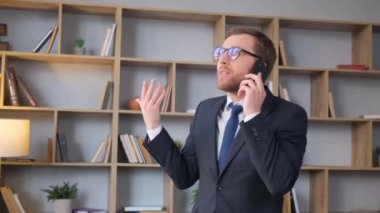 Arka planda kitap rafları olan modern bir ofiste dururken müşterilerle telefonda konuşmakla meşgul olumlu bir yönetici. İşte meşgul genç bir şirket müdürü.
