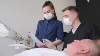 Diş muayenehanesindeki kıdemli bir adam dişlerini genç bir dişçi ve bayan bir asistanın yardımıyla kontrol ettiriyor.. 