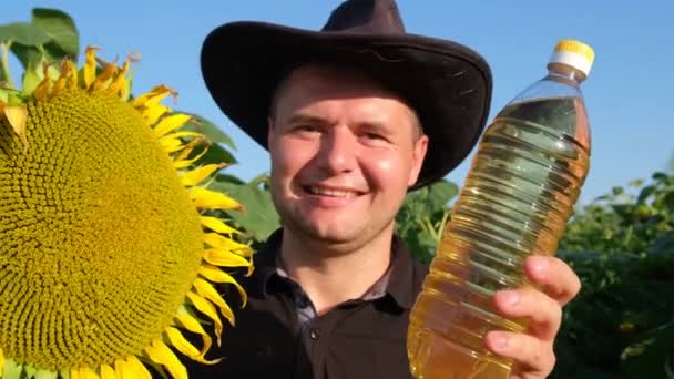 快乐的农民在向日葵地里拿着一瓶油 一个年轻的农民在田里抱着一个成熟的向日葵头 — 图库视频影像