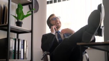 A happy businessman at the workplace has put his feet up on the table and is talking on a smartphone. Confident and defiant office worker behind a laptop put his feet on the table. Vertical video