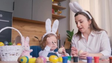 Anne ve kızı Paskalya için yumurta boyuyor. Mutlu bir aile tatil için hazırlanıyor. Tavşan kulağı takan tatlı küçük bir kız. 4k video