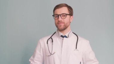 Gri arka planda gözlüklü genç bir doktorun portresi, kameranın merceğine bakar ve elini sallar. İnsanları tedavi etme kavramı. Dikey video
