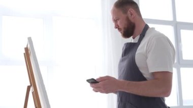 Yetenekli bir erkek sanatçı, fırça kullanarak, beyaz bir tuvalde yağlı boya tablosunun modern bir başyapıtını yaratıyor. Tuval büyük bir stüdyoda bir sehpanın üzerinde..