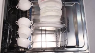 Bir kadın lastik eldiven verip, beyaz tabak ve bardakları bulaşık makinesine koyar. İleri teknolojileri kullanarak yüksek kaliteli mutfak gereçleri.
