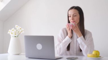 Dizüstü bilgisayarda çalışan güzel genç bir kadın evde oturup kahve içiyor. Merkez ofis kavramı.